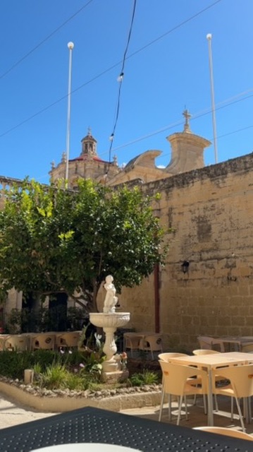 The shaded courtyard with a stone fountain and the parish church dome behind